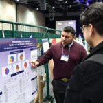 A man points at a research poster while a second man wearing a backpack listens.