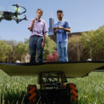 Two men stand on a grass field with the Willis Tower in the background. In the foreground, an aerial drone flies towards a small remote-controlled vehicle with a flat square on its roof.