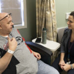 A man in a sweatshirt with glasses on his foreheads talks with a woman in a gray suit in a medical clinic examination room.