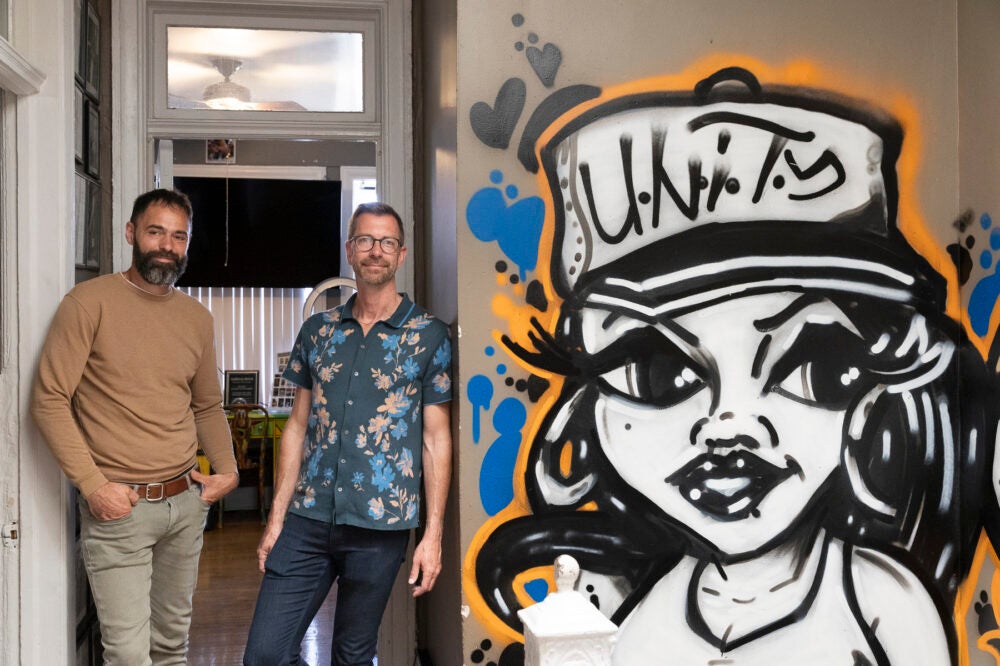 Two men stand in a doorway next to a graffiti painting of a woman wearing a baseball cap with the word "unity"