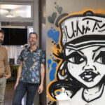 Two men stand in a doorway next to a graffiti painting of a woman wearing a baseball cap with the word "unity"