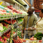 Young African American couple of shoppers standing by large display with fresh vegetables and choosing tomatoes and other products