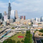 Aerial view of the University of Illinois Chicago campus, showcasing a large gathering of people in red shirts on a field near campus. The cityscape of Chicago is visible in the background.