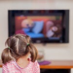 A little girl with pigtails seen from behind watching a flat-screen TV that is out of focus in the background.