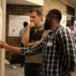 A man with his hand on his chin looks at a poster while a second man points at it.