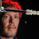 A slightly out-of-focus man stands behind black and white laboratory piping with a single drop of water falling.