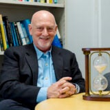 A man in a suit jacket and blue collared shirt sits at a table next to a brown wooden hourglass in front of a bookcase.