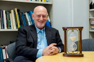 A man in a suit jacket and blue collared shirt sits at a table next to a brown wooden hourglass in front of a bookcase.