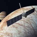 A space craft with two wide rectangular panels hovers above a white and brown moon with the planet Jupiter in the background.