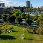 A drone photo of a green field with plants and a tall metal tower in the foreground and campus buildings in the background.