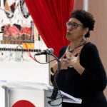 A woman wearing a black dress and glasses speaks at a lectern