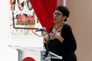 A woman wearing a black dress and glasses speaks at a lectern