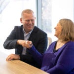 A man in suit jacket and a women in a purple shirt bump fists together.