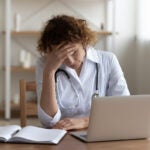 A woman in a white doctor coat with a stethoscope around her neck holds her hand to her forehead while looking at a laptop.