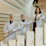 A man and two women in lab coats stand on a white concrete spiral staircase, leaning on a silver railing.