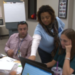 A standing woman in a blue shirt points at a laptop screen as a man and a woman on either side of her look at the screen.