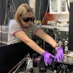 A woman in dark glasses and a striped shirt works on scientific equipment in a laboratory.