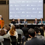 Q&A discussion led by a woman standing at a podium with six panelists seated at a table.