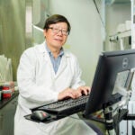 A man in a white lab coat and glasses sits at a computer in a lab
