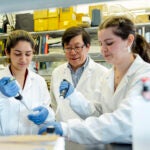 A professor supervises as two students work with samples in a lab