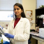 A woman in a white lab coat and blue gloves holds a pipette while working at a lab bench.
