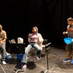 Three students stand in a dark rehearsal room behind props of music stands. A man in a beanie is on the left, a woman with long, purple hair is in the middle and a man on the right is holding a blue bowl and a beer.