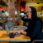 A woman smiles while sitting at a computer in a cafe.